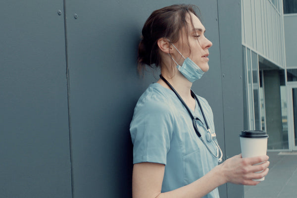 Tired and burned out nurse leaning up against wall with disheveled hair and drinking coffee