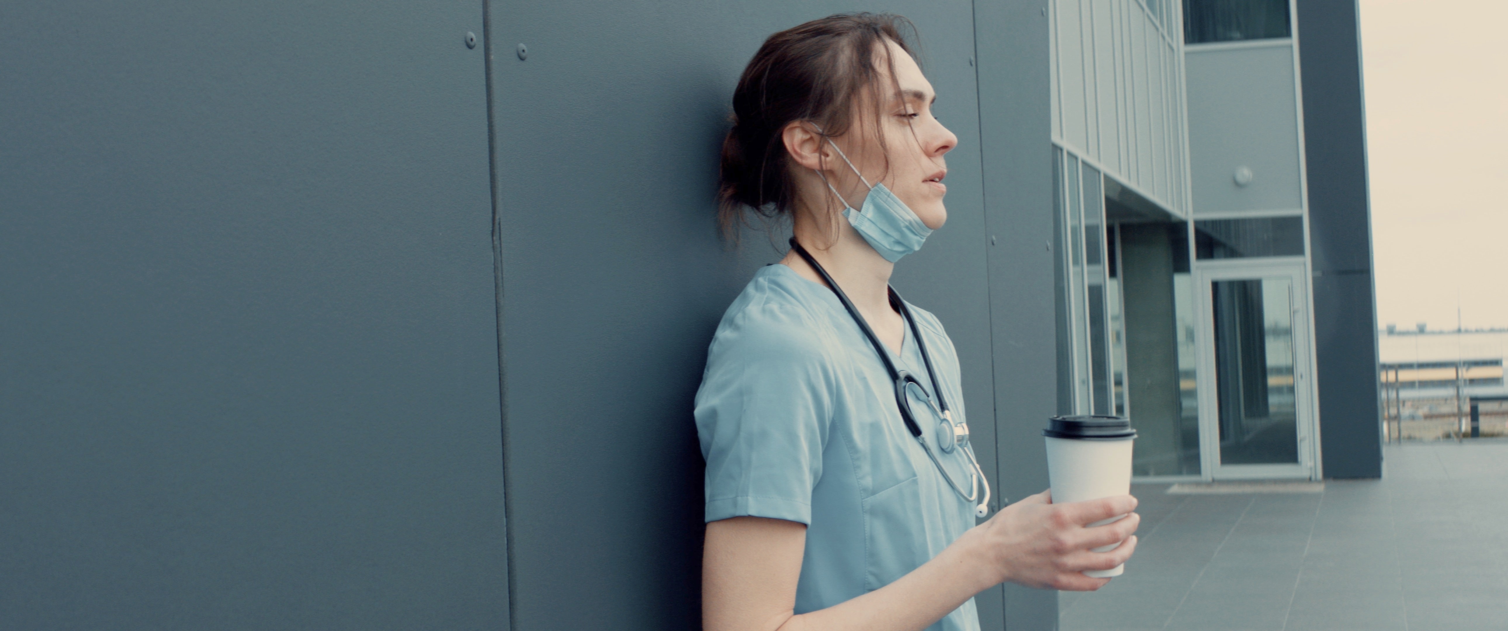 Tired and burned out nurse leaning up against wall with disheveled hair and drinking coffee